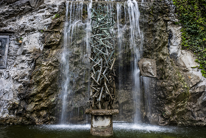 Scultura colonnare in metallo su piedistallo nella cascata di una fontana incastonata nella parete rocciosa di Ivrea