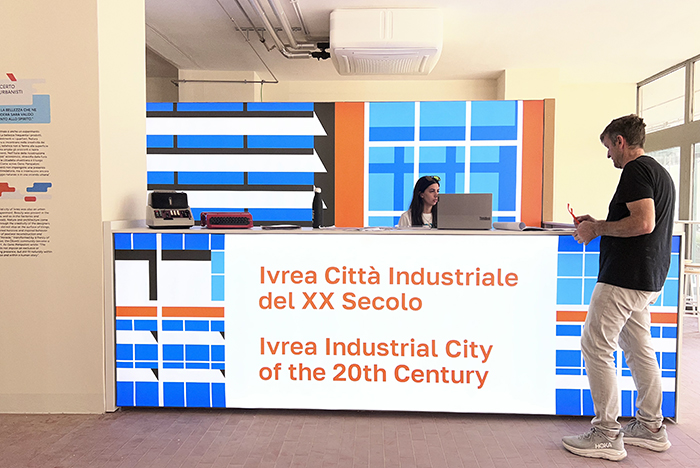 Banco accoglienza mostra 'Ivrea Città Industriale del XX Secolo' con operatrice e visitatore in centro espositivo di Ivrea