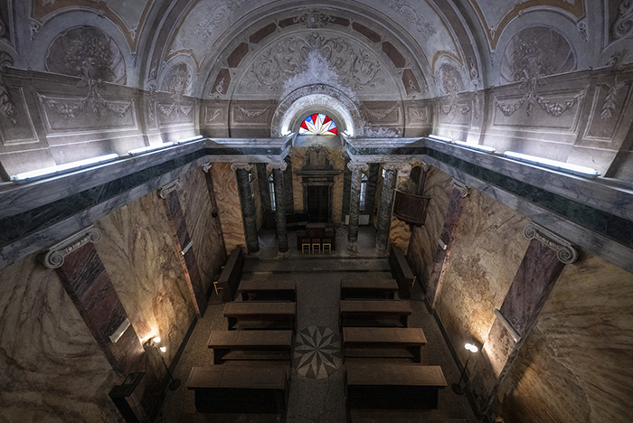 Interno di cappella storica a Ivrea con banchi in legno, colonne marmoree e abside con vetrata