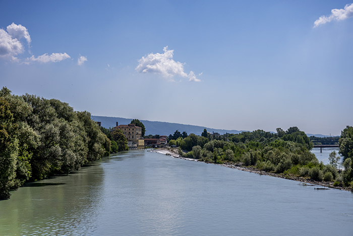 Calm river flowing between tree-lined green banks toward distant buildings and low hills under a blue sky