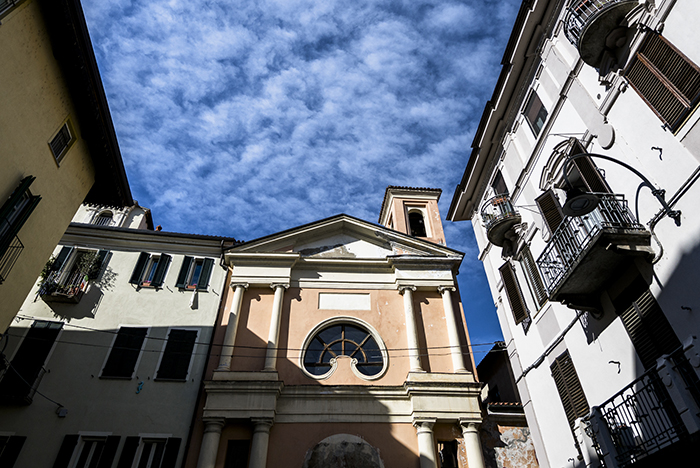 Peach-colored church facade with round window and bell tower flanked by buildings under a blue sky with wispy clouds
