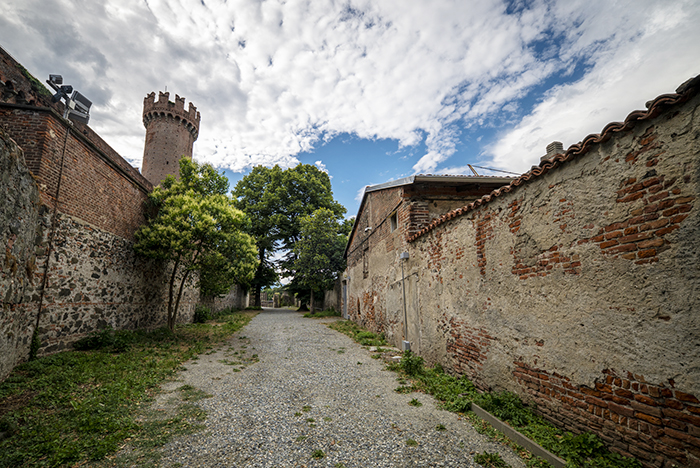 Gravel lane between weathered brick and stone walls leading to trees and a round crenellated tower under a cloudy blue sky